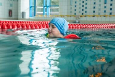 Kind mit blauer Badekappe schwimmt mit Schwimmflügeln im Becken des Rolli-Bades