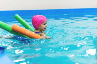 kleines Mädchen mit pinker Badekappe lernt mit Poolnudel schwimmen